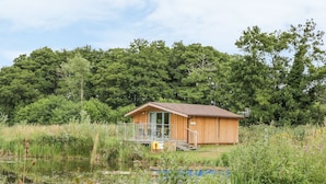 A wooden cabin with a glass door, situated by a pond surrounded by lush greenery. - Lakeside Lodge (Norwich)