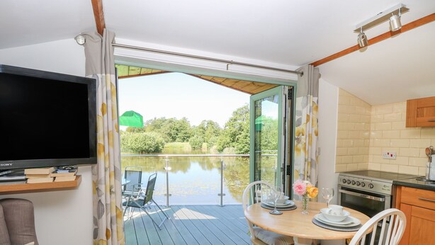 A room with a TV, a dining table with chairs, a kitchen area, and a large window overlooking a body of water. - Lakeside Lodge (Norwich)