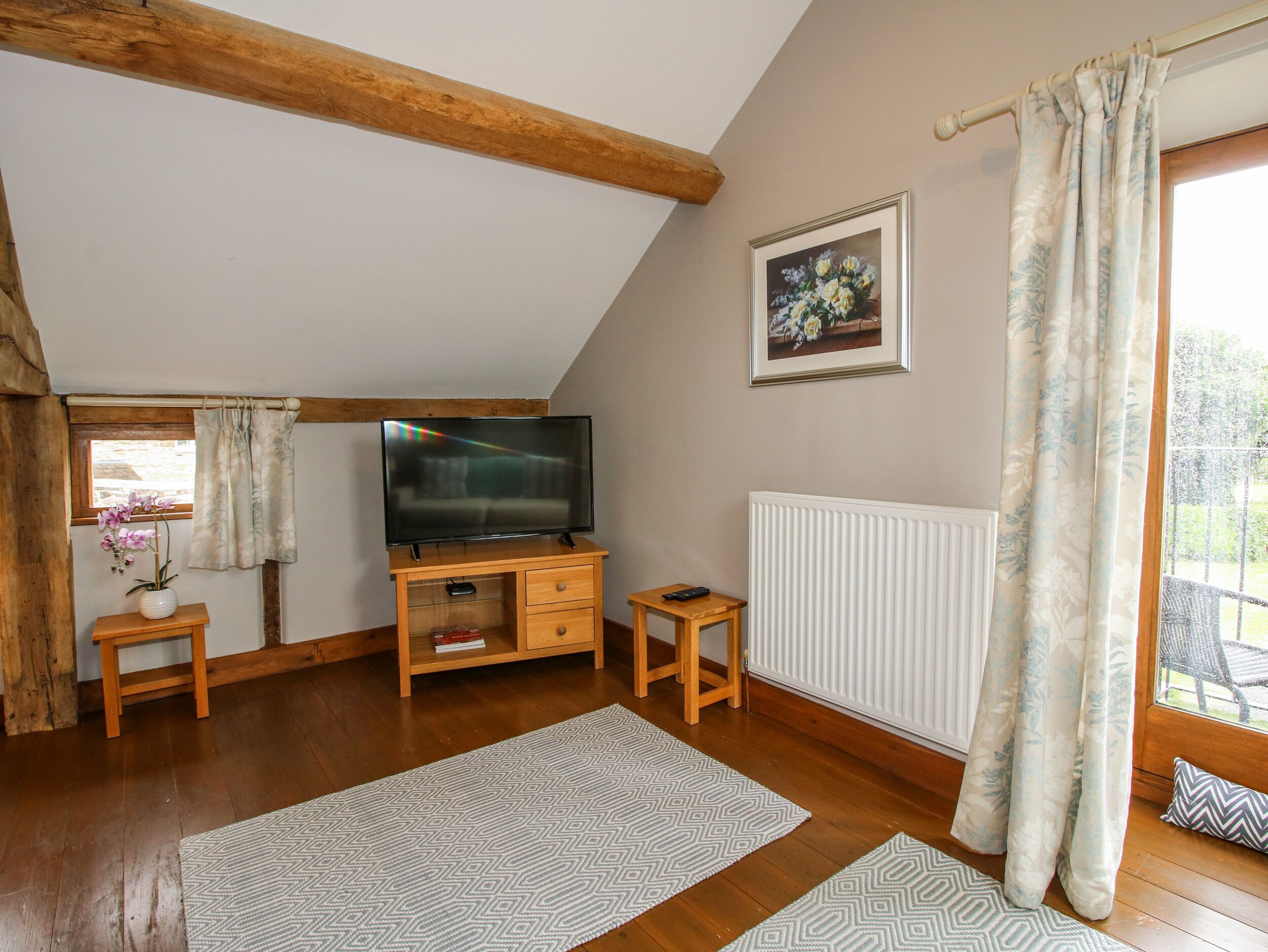 A cozy room with a sloped ceiling, wooden beams, a television on a wooden stand, a framed picture on the wall, and a window with curtains.