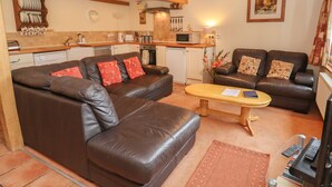 A living room with a brown leather sofa, a wooden coffee table, and a kitchen area with white cabinets and a microwave. - The Old Stables (Ross-on-Wye)