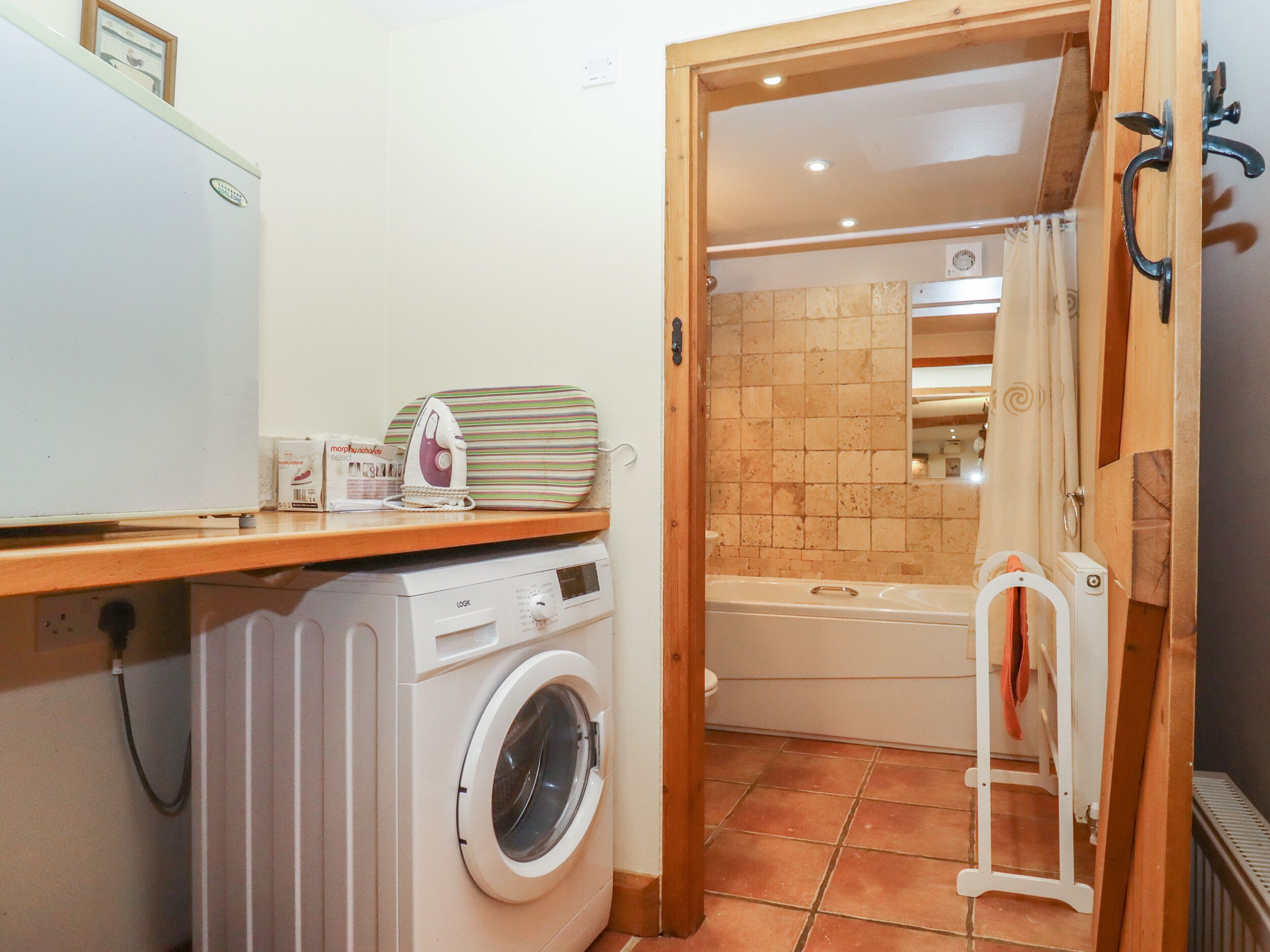 A compact laundry room with a washing machine, an ironing board, and a bathtub.