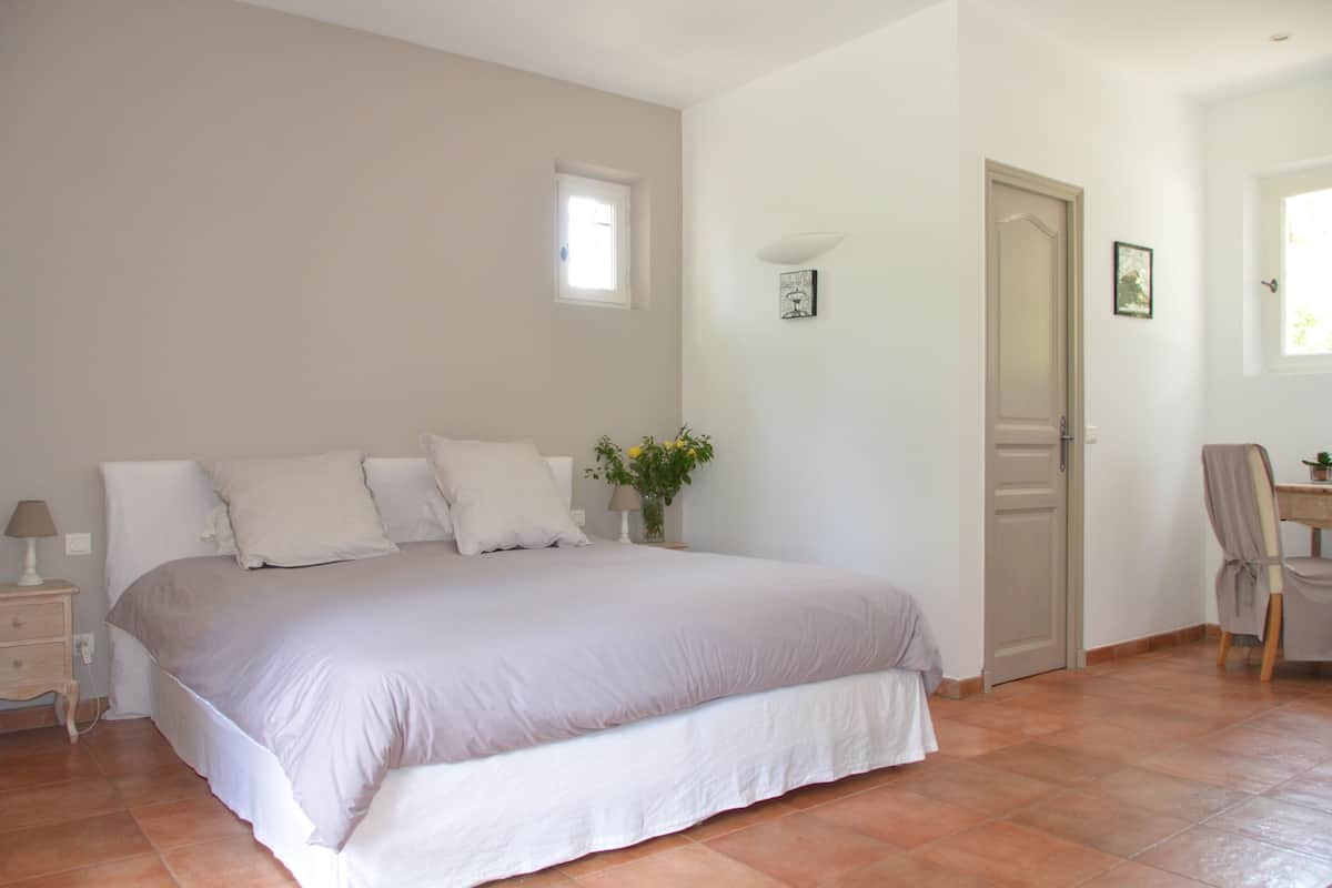Ein Schlafzimmer mit einem großen Bett, einem Nachttisch, einem Sessel, einer Tür und einem kleinen Fenster.