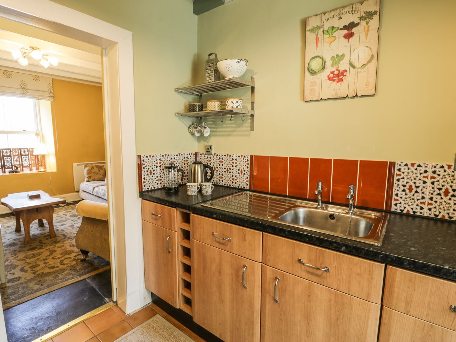 Cocina con armarios de madera, encimera negra y un lavabo. Se ve una ventana que ofrece vistas a una habitación con sofá y mesa.