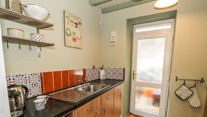 A kitchen with wooden cabinets, a black countertop, and a sink. There are shelves above the counter holding kitchenware. A door with a frosted glass panel is visible, and kitchen towels are hanging on a rack. - Miners Cottage (Blaenau Ffestiniog)