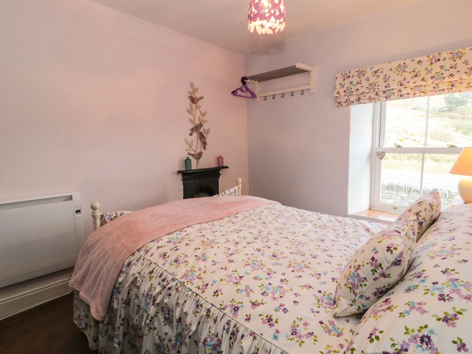 A bedroom with a floral bedspread, a radiator, a wall-mounted heater, a window with a floral valance, and a hanging light fixture.