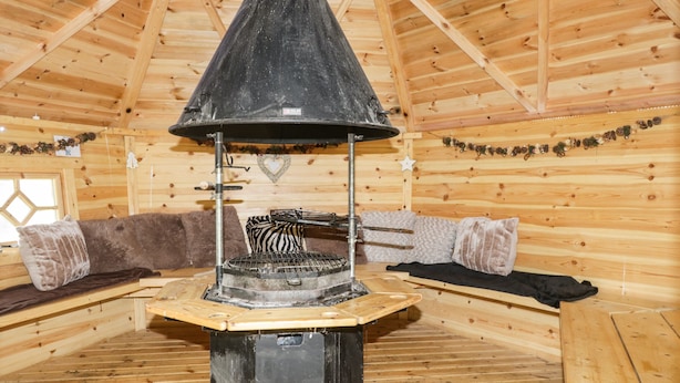 A wooden cabin interior with a central wood stove, wooden walls, and a cozy seating area. - The Cottage at Graysondale Farm (Cockermouth)