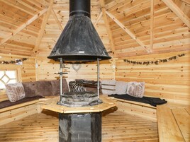 Interior de una cabaña de madera con un fogón central, paredes de madera y una zona de estar acogedora.