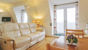 Una sala de estar con un sofá color beige, una mesa de comedor de madera con sillas, una mesita auxiliar con una lámpara y un balcón con cortinas florales.