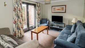 A living room with a sofa, a coffee table, a television on a stand, and a window with curtains. - The Lodge off High Street (Ipswich)
