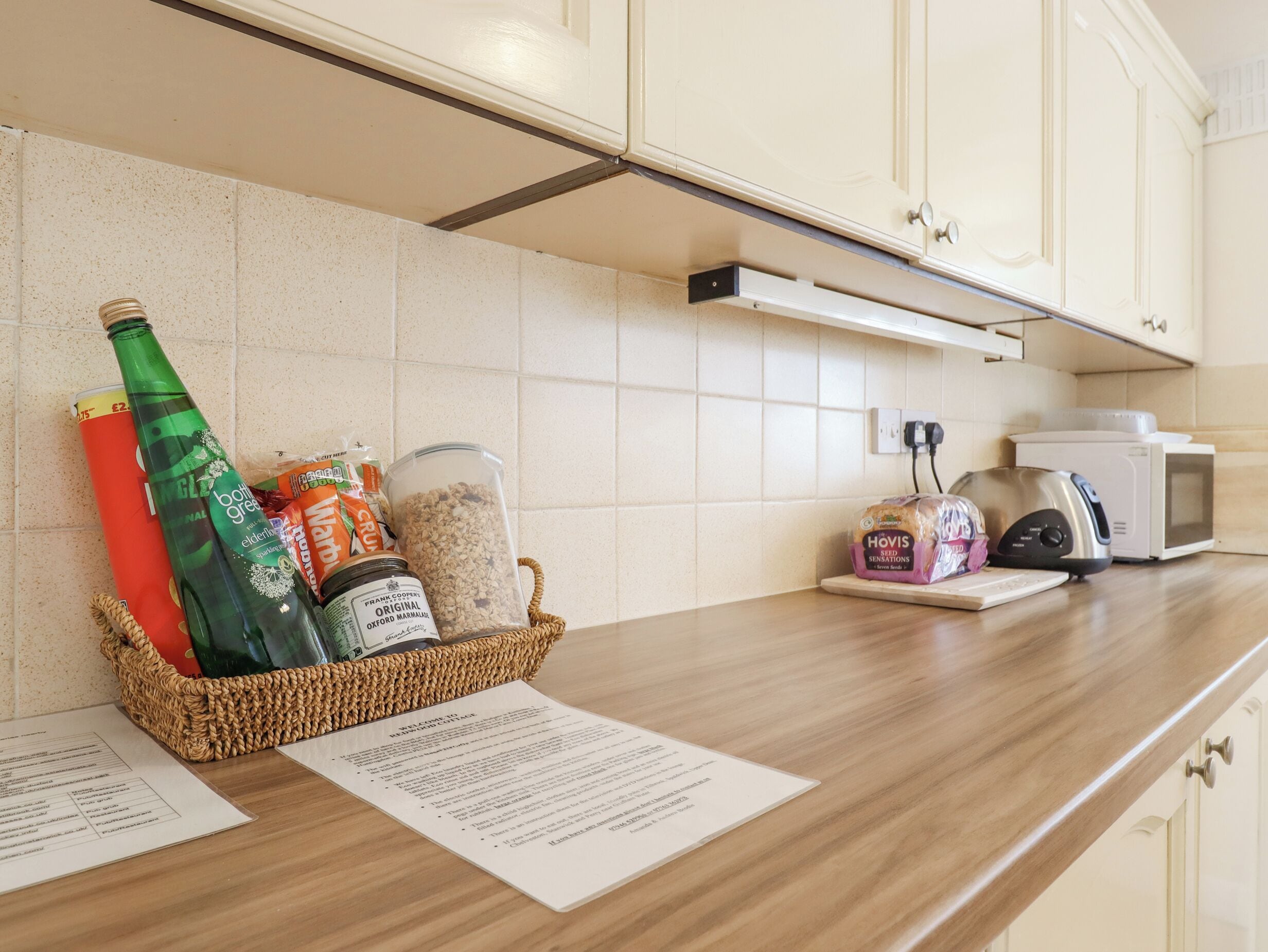 A kitchen counter with a basket containing a green bottle, a red bottle, and various food items. There is a microwave and a toaster on the counter.