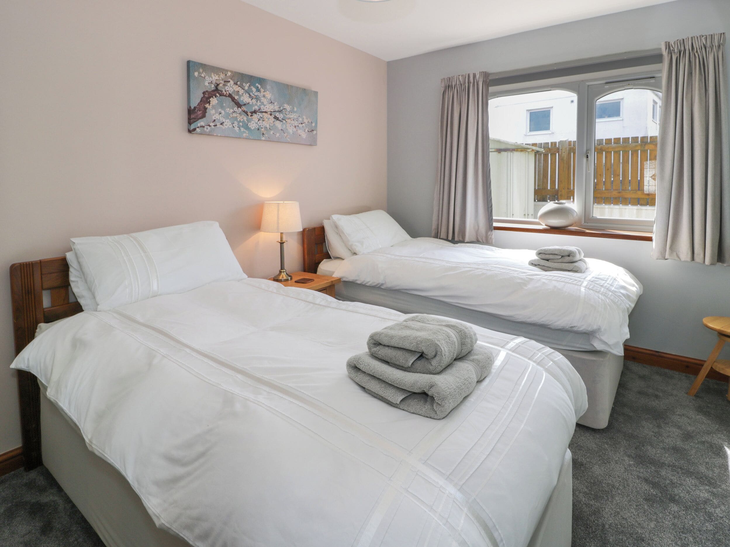 Quarto de hotel com duas camas, uma mesa de cabeceira com luminária, um quadro na parede e uma janela com cortinas.