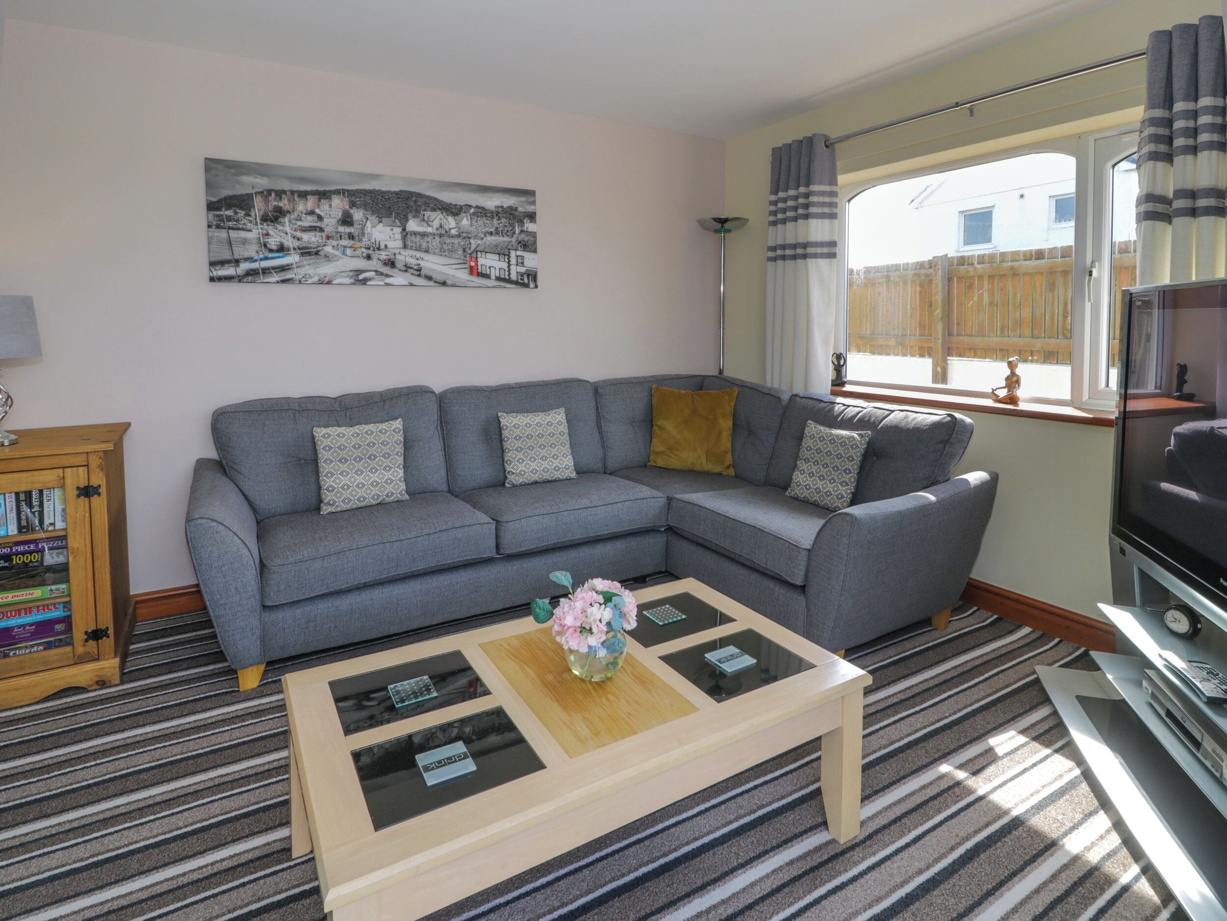A living room with a grey sofa, a wooden coffee table with a vase, a bookshelf, and a painting on the wall.