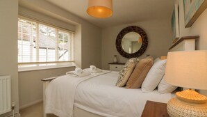 A bedroom with a bed, a bedside table, a lamp, a mirror, and a window with blinds. - Heron Cottage (Ilfracombe)