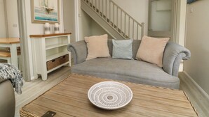 A living room with a grey sofa, a wooden coffee table with a white decorative bowl, a staircase, and a painting on the wall. - Heron Cottage (Ilfracombe)