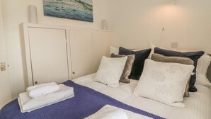 A neatly made bed with white and blue bedding, a painting of a coastal scene above it, and a wall-mounted light fixture. - Wren Cottage (Ilfracombe)