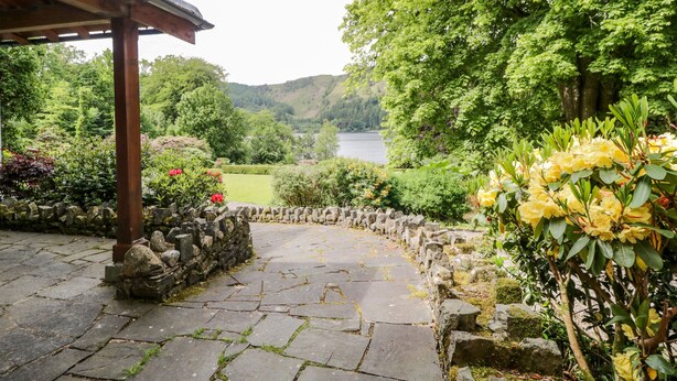 A stone pathway leading to a wooden pergola, surrounded by lush greenery and a view of a lake. - Thirlmere Suite (Keswick)