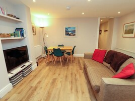 A living room with a TV, wooden flooring, a dining table with chairs, a sofa with red cushions, and a painting on the wall.