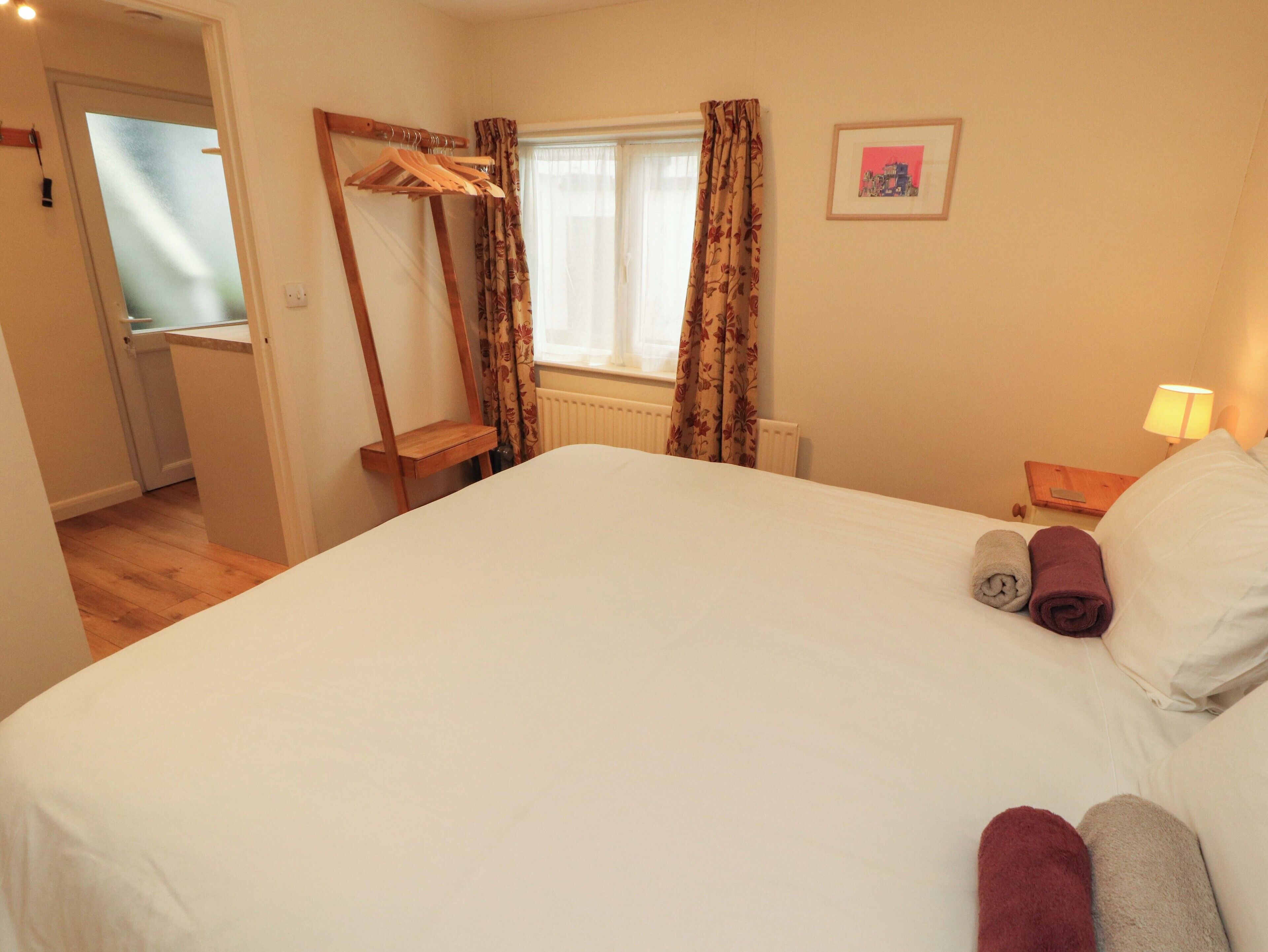 Ein Schlafzimmer mit einem Bett, einem Kleiderhaken aus Holz, einem Fenster mit Vorhängen, einem Nachttisch mit Lampe und einem gerahmten Bild an der Wand.