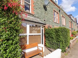 A traditional stone house with a wooden bench outside, a brick wall, and a hedge.