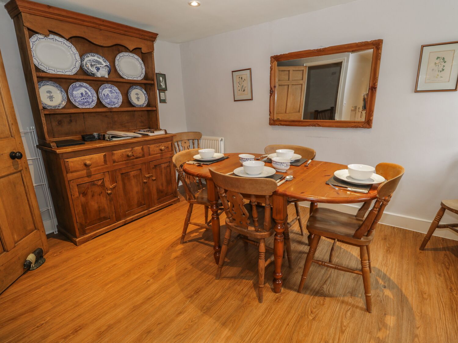 Un comedor con una mesa de madera dispuesta para cuatro personas, rodeada de sillas de madera. A la izquierda, un aparador de madera con platos azules y blancos.