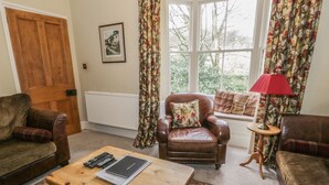 A cozy living room with a brown leather armchair, a wooden coffee table with remote controls and a book, and a window with floral curtains. - Rowan Cottage (Ambleside)