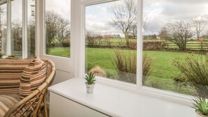 A room with large windows overlooking a grassy field and trees. There is a wicker chair and a small potted plant on a windowsill. - Low Garth Cottage (Penrith)