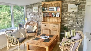 A cozy room with wicker furniture, a wooden table, and a bookshelf against a stone wall. - Low Garth Cottage (Penrith)