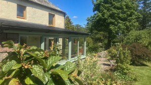 A house with a conservatory, surrounded by a garden with various plants and a large tree. - Low Garth Cottage (Penrith)