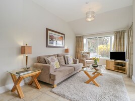 A living room with a beige sofa, a glass coffee table, a TV on a wooden stand, and a large window with curtains.