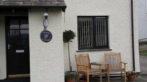 Une petite maison avec une pancarte indiquant « Thyme Cottage », une porte noire, une fenêtre avec des barreaux verticaux, et deux chaises en bois et une table à l’extérieur.