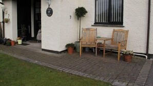 Une maison à l’extérieur blanc, avec une véranda en bois dotée de deux chaises, un pot de fleurs et une fenêtre avec des stores verticaux.