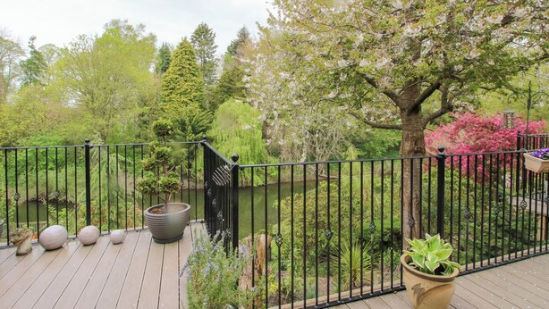 A wooden deck with a metal railing, surrounded by various potted plants and trees. - Riverdale (Shrewsbury)