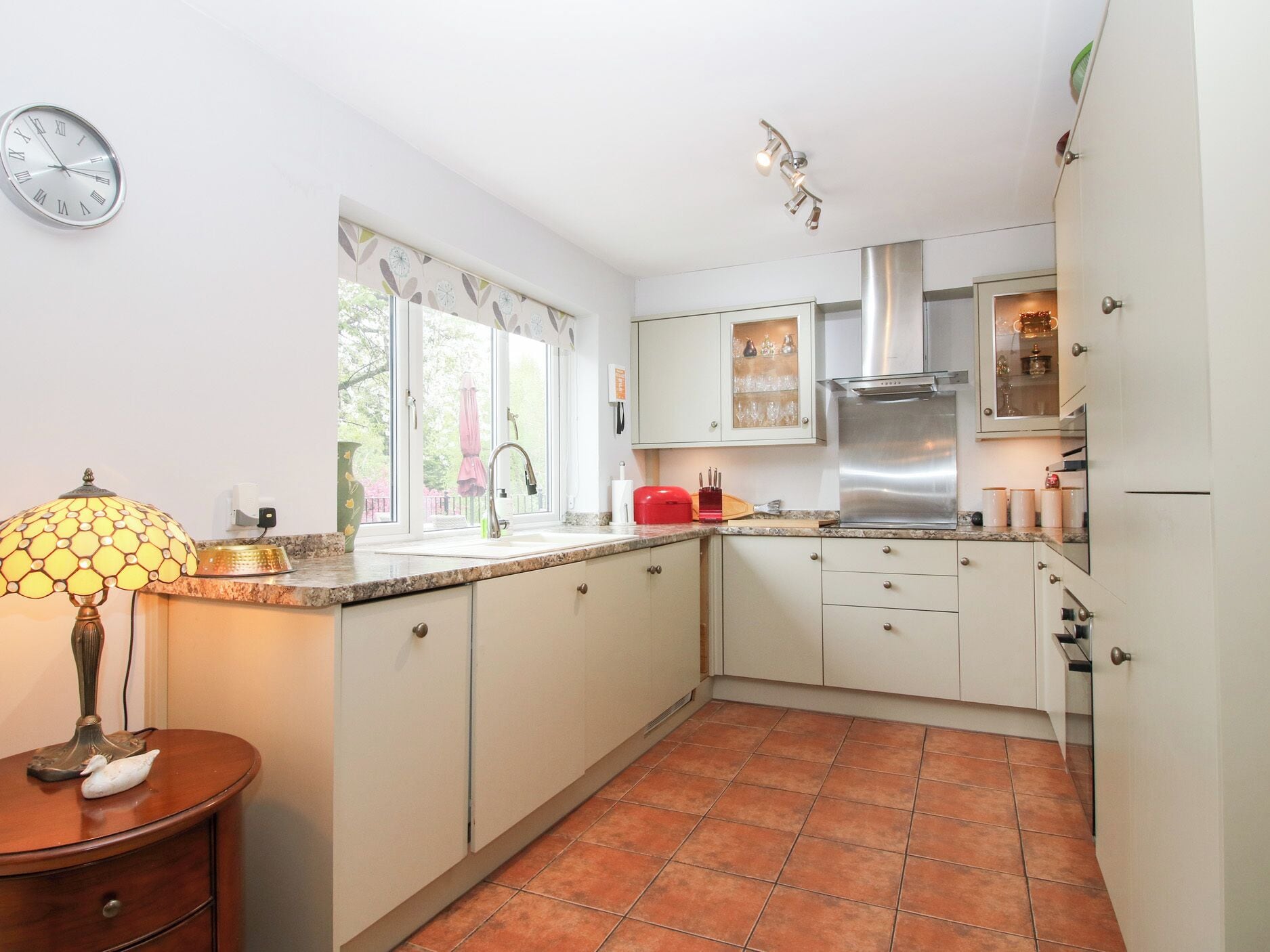 A kitchen with white cabinets, a stainless steel range hood, a window with a view, and a clock on the wall.