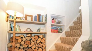 Un escalier recouvert d’une moquette à rayures, une pile de bois de chauffage et des étagères remplies de livres et de jeux de société.