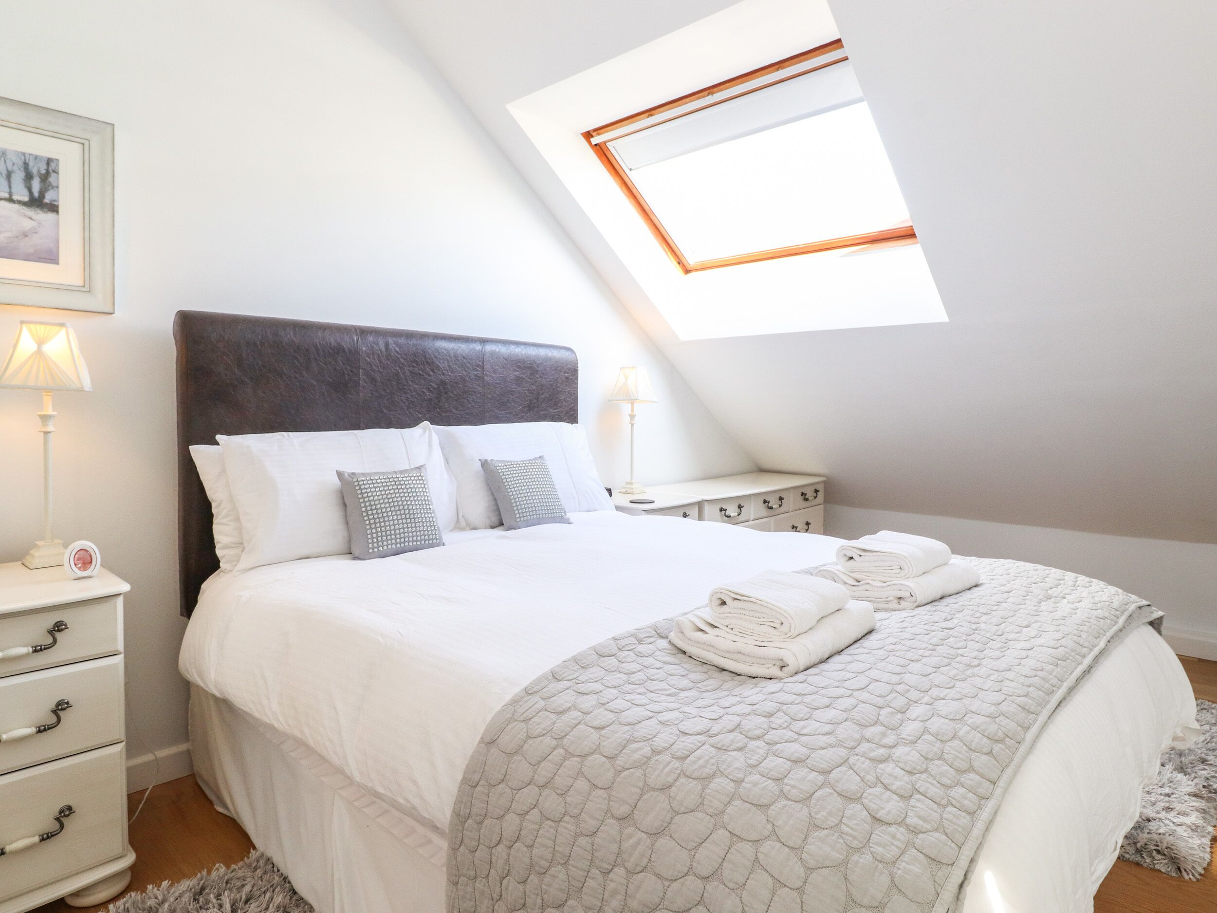 A bedroom with a bed, two bedside tables, a nightstand, a window, and a framed picture on the wall.