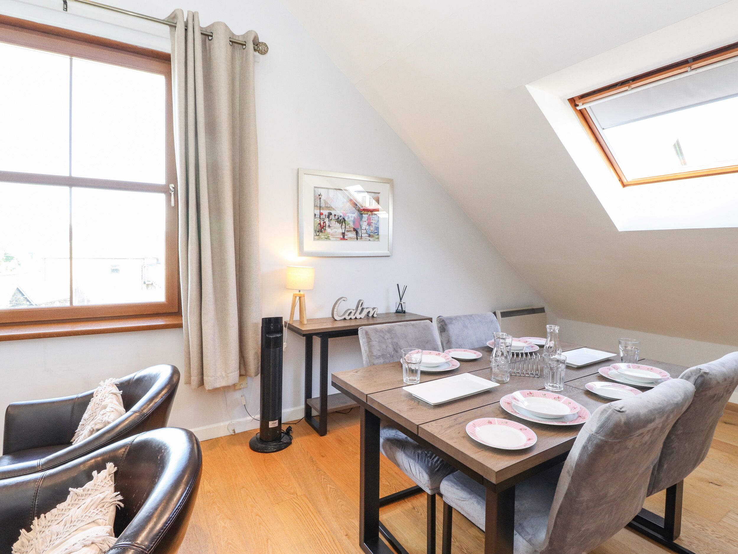 A dining area with a table set for four, a standing fan, and a window with curtains.