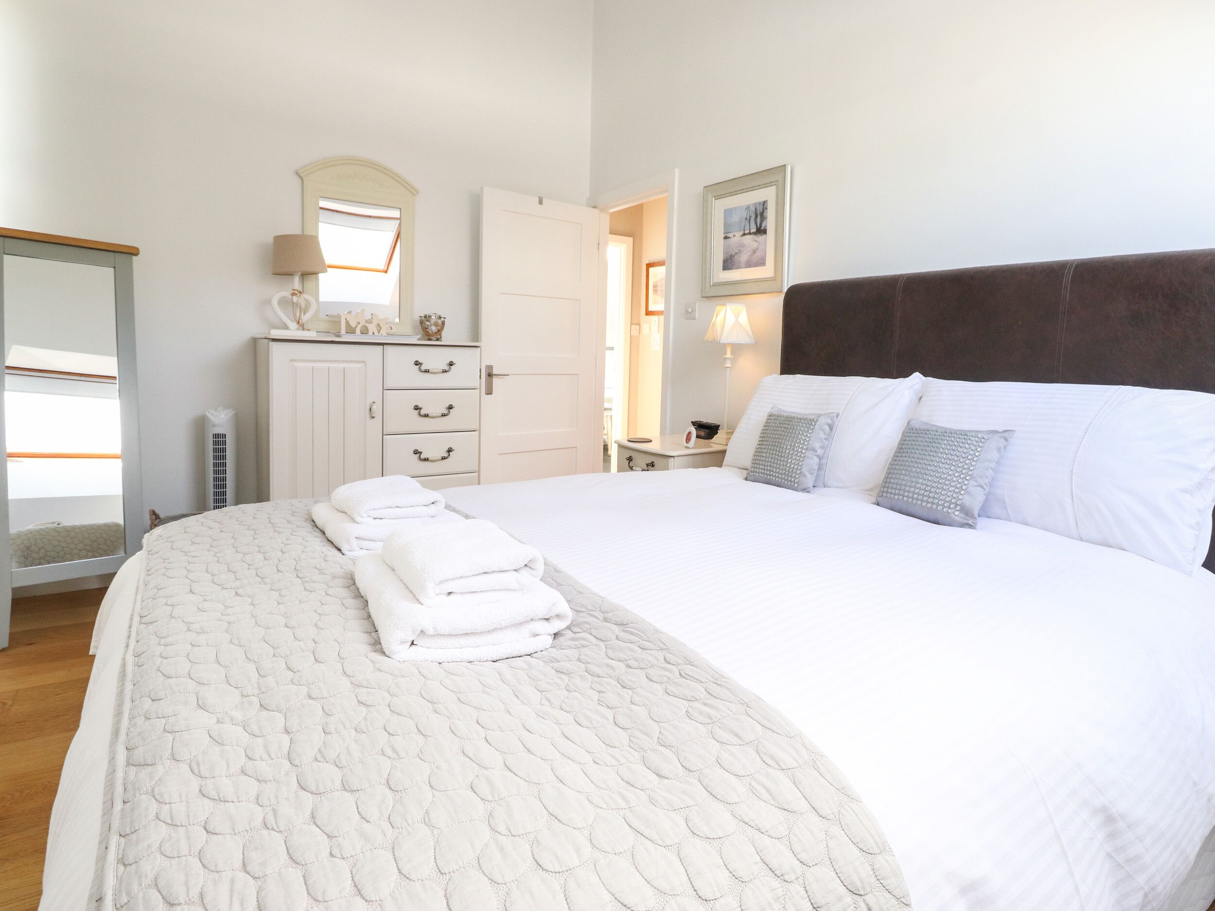 A neatly made bed with white linens and pillows, a dresser with drawers, a mirror, and a framed picture on the wall.