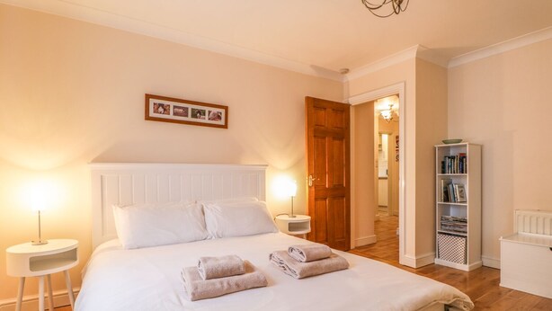 Een net opgemaakt bed met witte lakens en twee gevouwen handdoeken, een nachtkastje met een lamp, een houten deur en een boekenplank met boeken.