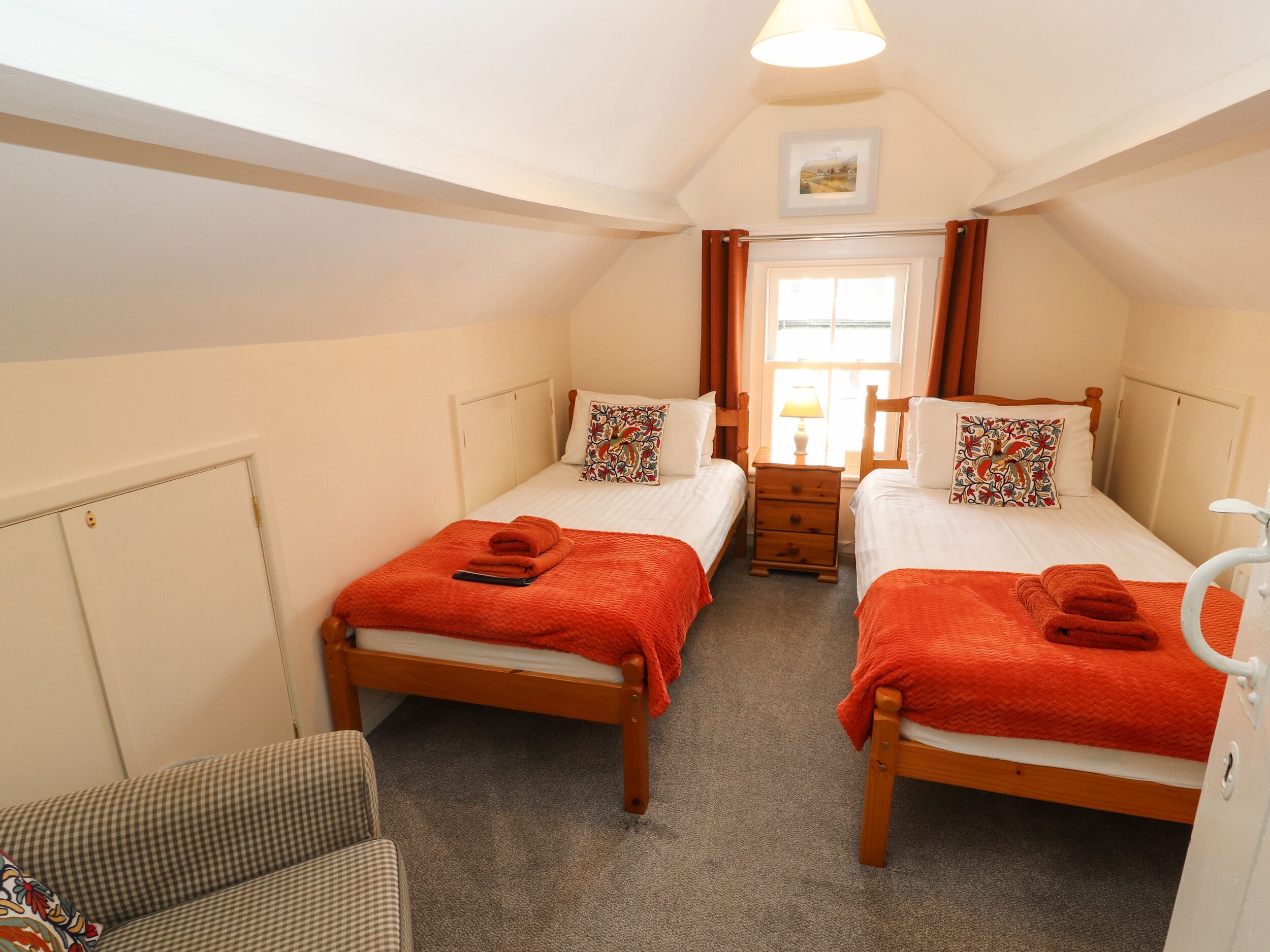 A small, cozy bedroom with two single beds, a wooden dresser, a chair, and a window with curtains.