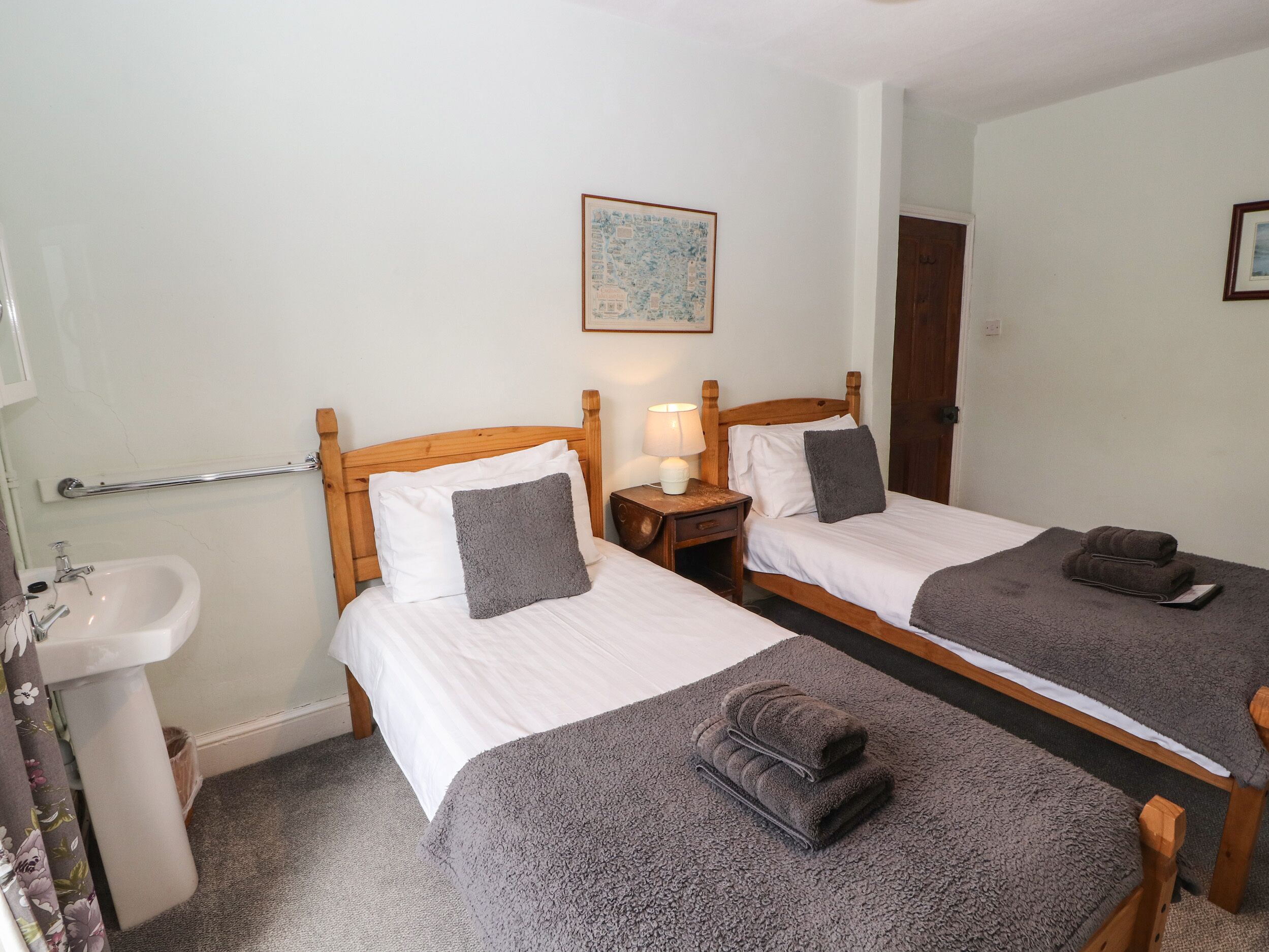 A hotel room with two single beds, a wooden headboard, a bedside table with a lamp, a small bedside table with a lamp, a mirror, a wooden door, a white sink, and a framed picture on the wall.