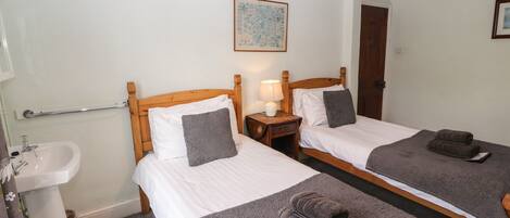 A hotel room with two single beds, a wooden headboard, a bedside table with a lamp, a small bedside table with a lamp, a mirror, a wooden door, a white sink, and a framed picture on the wall.
