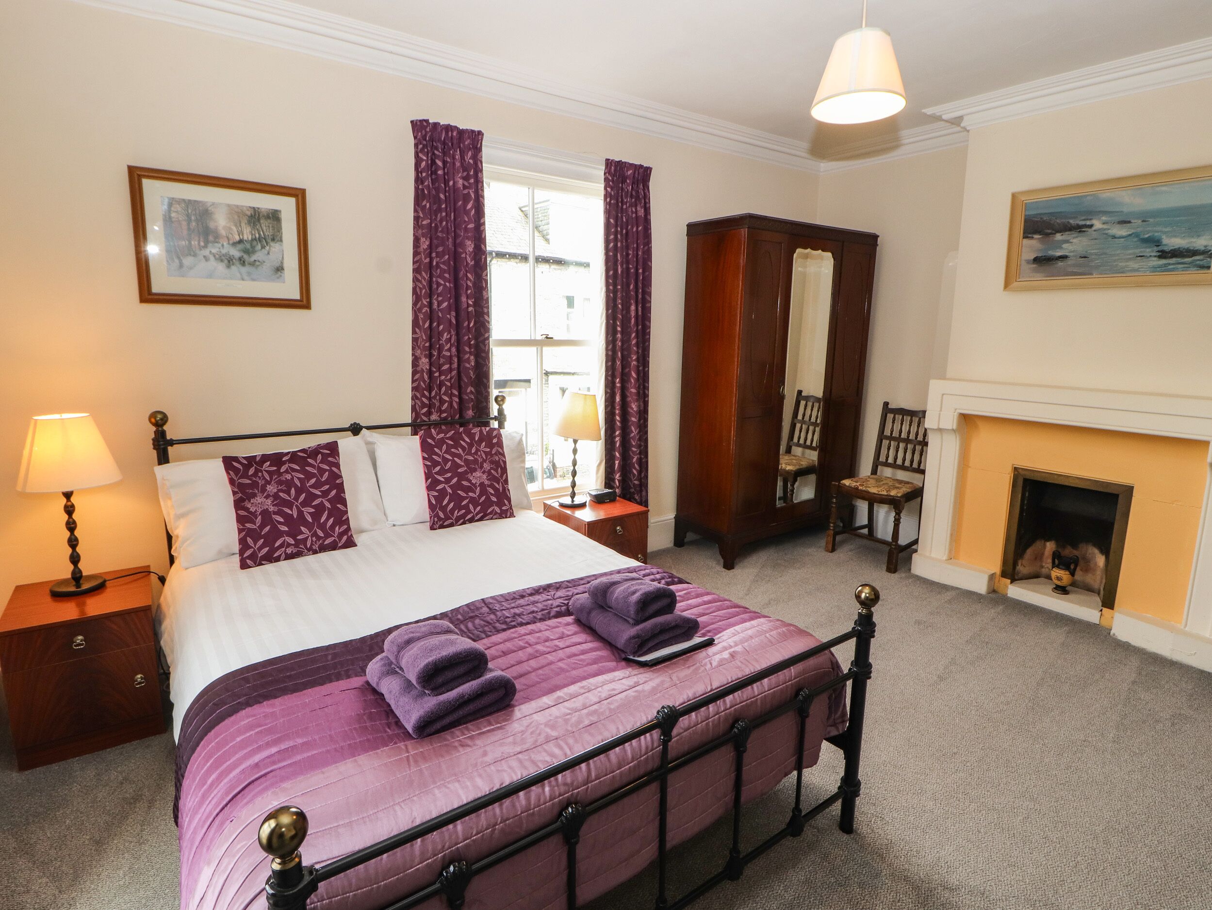A neatly arranged bedroom with a bed, bedside tables, a wardrobe, a chair, a fireplace, and a window with curtains.