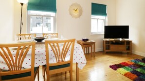 Un comedor con una mesa dispuesta para cuatro personas, un televisor sobre un mueble bajo y un reloj en la pared.
