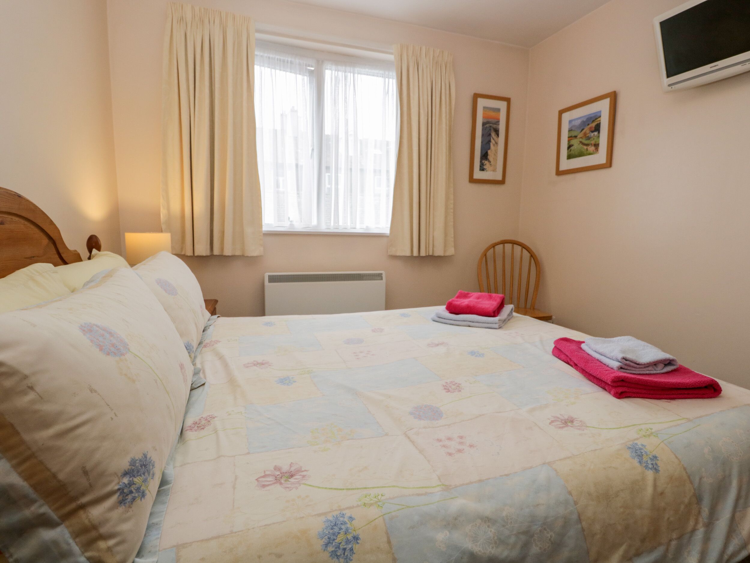 A bedroom with a bed, a chair, a window with curtains, a radiator, and two framed pictures on the wall.