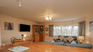 A cozy living room with a wooden dining table, a sofa with cushions, a bookshelf, and a television on the wall. - Treetops (Windermere)