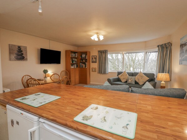 A cozy living room with a wooden dining table, a sofa with cushions, a bookshelf, and a television on the wall.