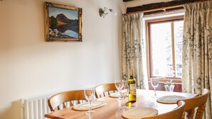 A dining table with chairs, a bottle of wine, and glasses. There is a painting on the wall and a window with curtains. - The Old Barn (Seascale)