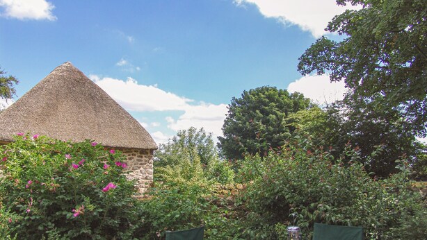 A round wooden table with a tray of tea set, surrounded by chairs, in a garden with a thatched-roof building and lush greenery. - The Bakehouse (Newton Abbot)