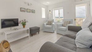 A modern living room with a flat-screen TV, a white console table, two grey sofas, a small armchair, and a large window offering a view of the landscape. - 1 The Quay (Exmouth)