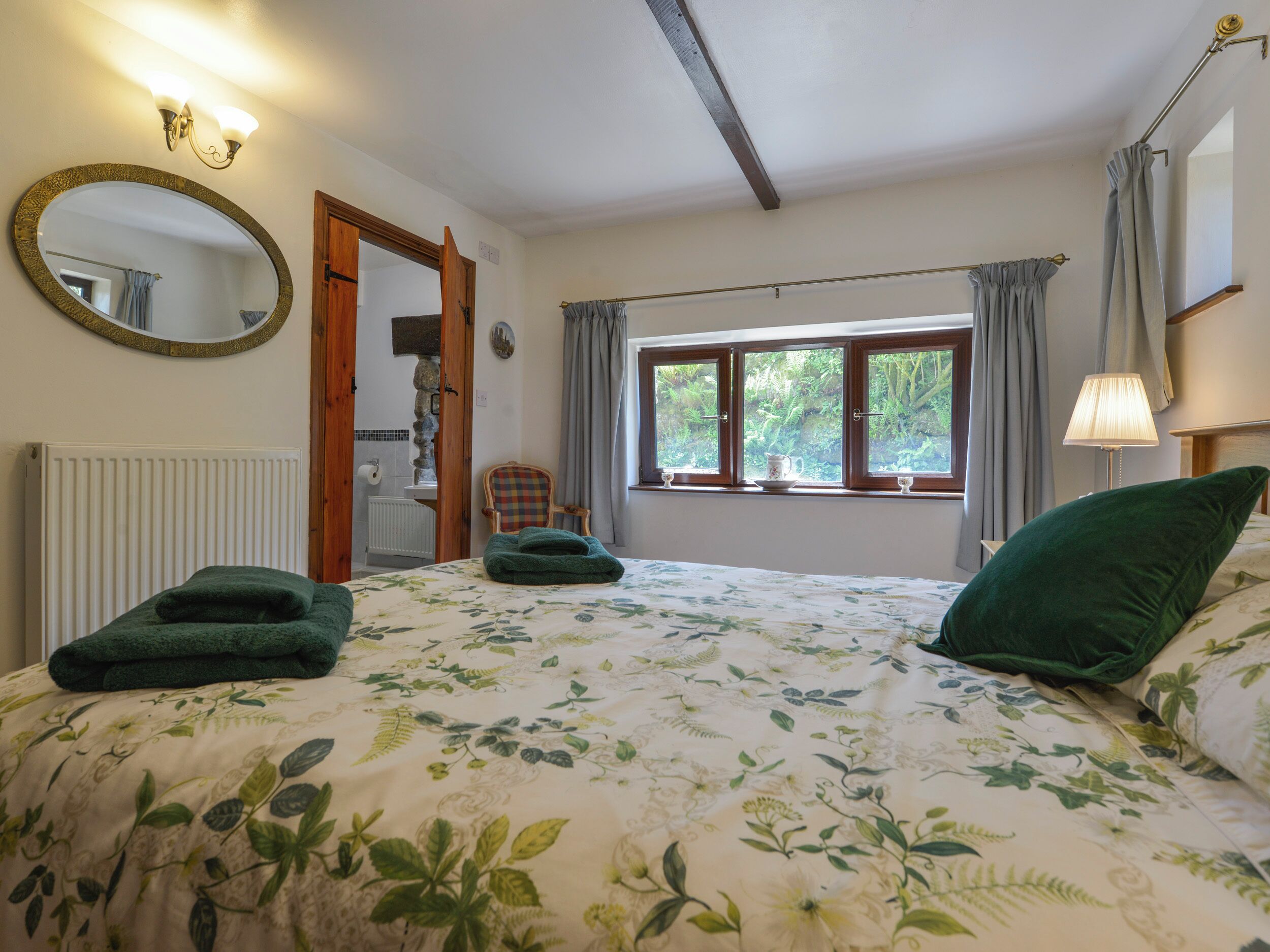A bedroom with a floral bedspread, a large mirror, a window with curtains, a radiator, and a chair.